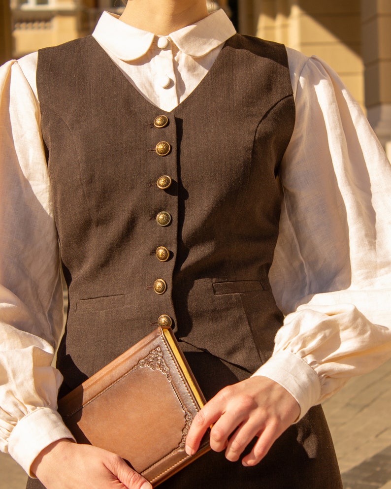 May include: A brown vest with gold buttons worn over a white collared shirt with puffed sleeves. The person is holding a brown leather-bound journal.