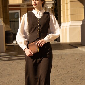 May include: A woman wearing a brown vest and skirt with a white blouse and a brown book in her hand.