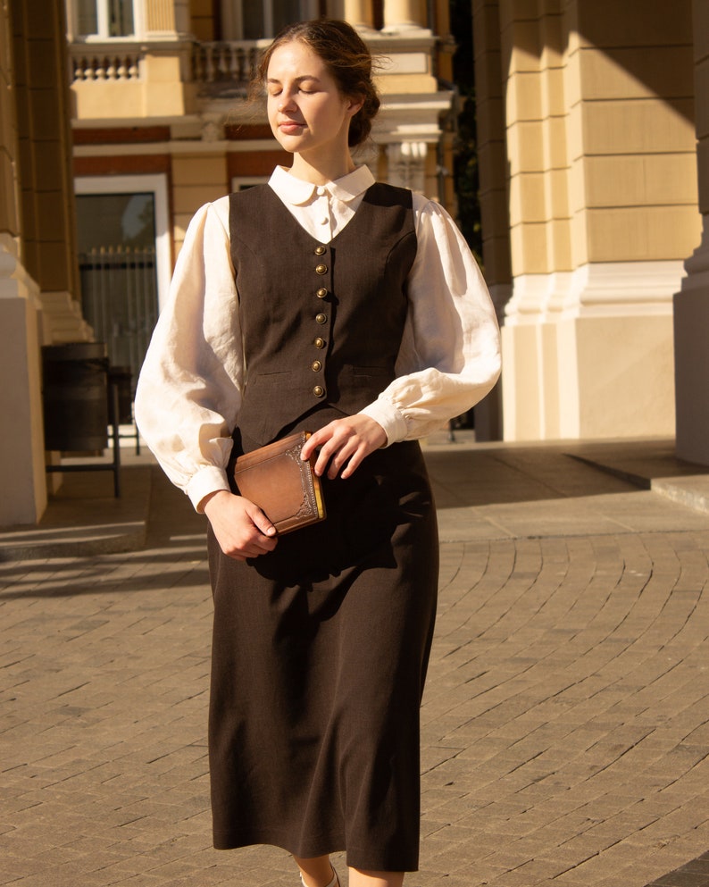 May include: A woman wearing a brown vest and skirt with a white blouse and a brown book in her hand.