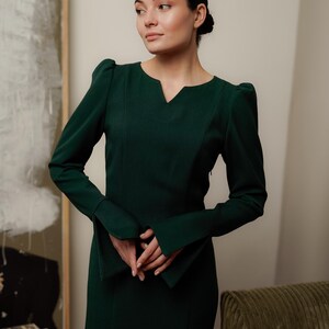 May include: A woman wearing a dark green long-sleeved dress with a V-neck and puffed sleeves. The dress has a fitted silhouette and a slightly flared skirt.