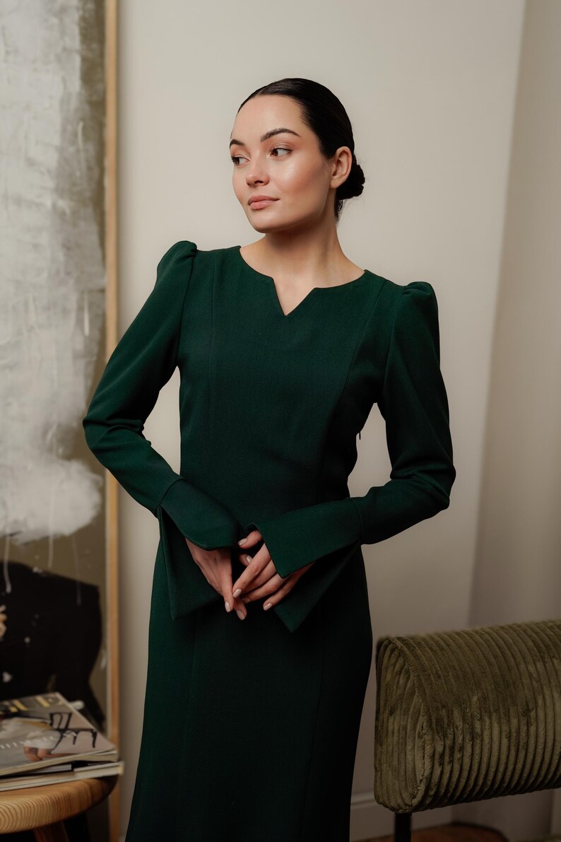May include: A woman wearing a dark green long-sleeved dress with a V-neck and puffed sleeves. The dress has a fitted silhouette and a slightly flared skirt.