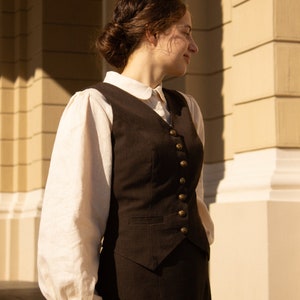 May include: A woman wearing a brown vest with gold buttons over a white blouse with a peter pan collar. She is standing in front of a building with a light brown facade.