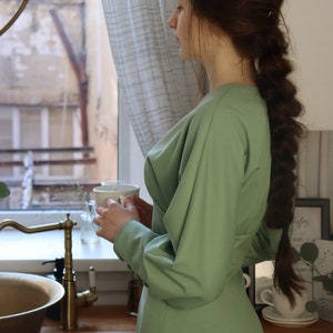 May include: A woman wearing a green dress stands in a kitchen, holding a white mug. She is looking out of the window. The kitchen has a wooden worktop, a sink, and a tap.