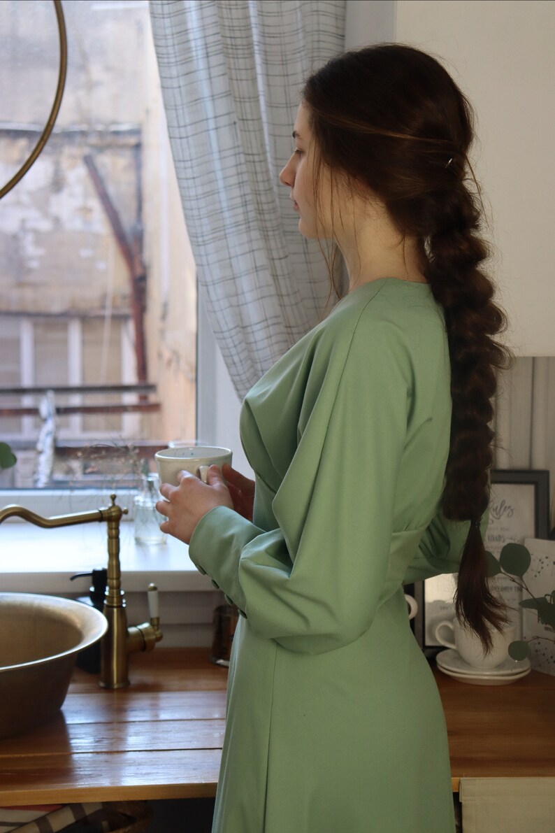 May include: A woman wearing a green dress stands in a kitchen, holding a white mug. She is looking out of the window. The kitchen has a wooden worktop, a sink, and a tap.