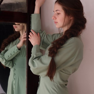 May include: A woman wearing a light green dress with long sleeves and buttons down the front. She has long brown hair braided down her back. She is standing in front of a mirror.