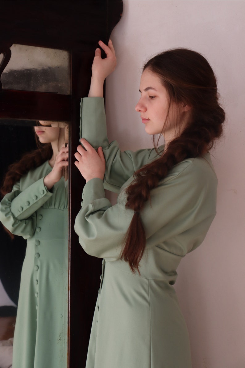 May include: A woman wearing a light green dress with long sleeves and buttons down the front. She has long brown hair braided down her back. She is standing in front of a mirror.