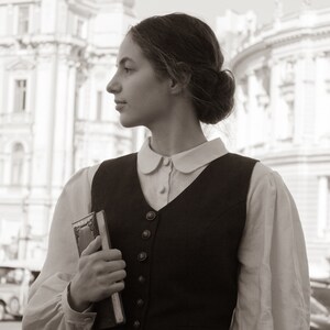 May include: A woman in a white blouse and a black vest with a white collar stands in front of a building. She is holding a closed book in her left hand.
