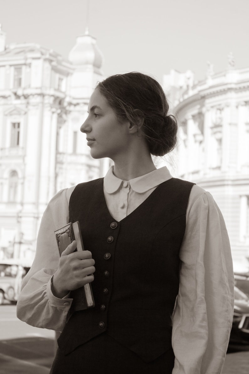 May include: A woman in a white blouse and a black vest with a white collar stands in front of a building. She is holding a closed book in her left hand.