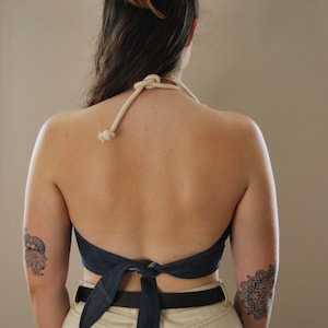 Puede incluir: Una mujer lleva un top de halter de mezclilla atado en un lazo en la espalda. El top se lleva sobre un pantal&oacute;n beige con un cintur&oacute;n negro. La mujer tiene un tatuaje en el brazo izquierdo.