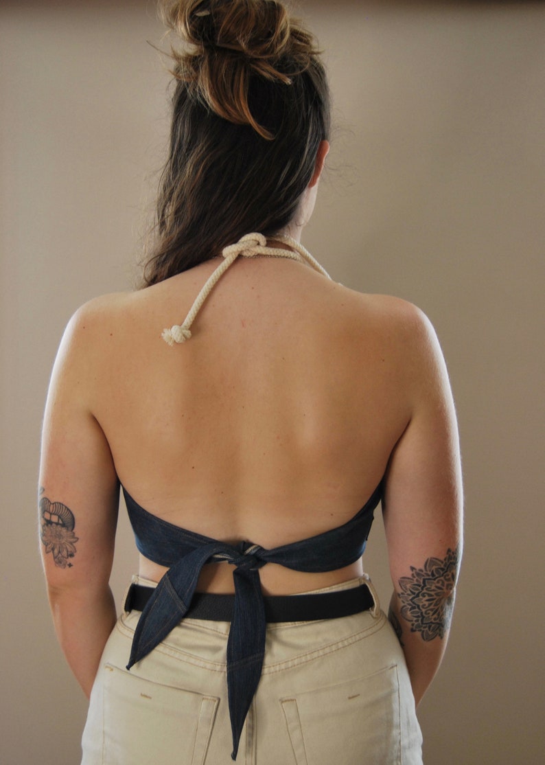 Puede incluir: Una mujer lleva un top de halter de mezclilla atado en un lazo en la espalda. El top se lleva sobre un pantal&oacute;n beige con un cintur&oacute;n negro. La mujer tiene un tatuaje en el brazo izquierdo.