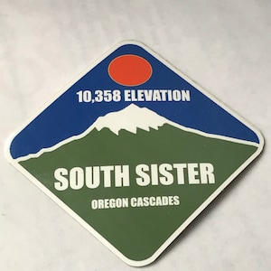 May include: A diamond-shaped sticker with a green, blue, and white mountain scene. The sticker says "South Sister" and "Oregon Cascades" with a red sun and the text "10,358 Elevation".