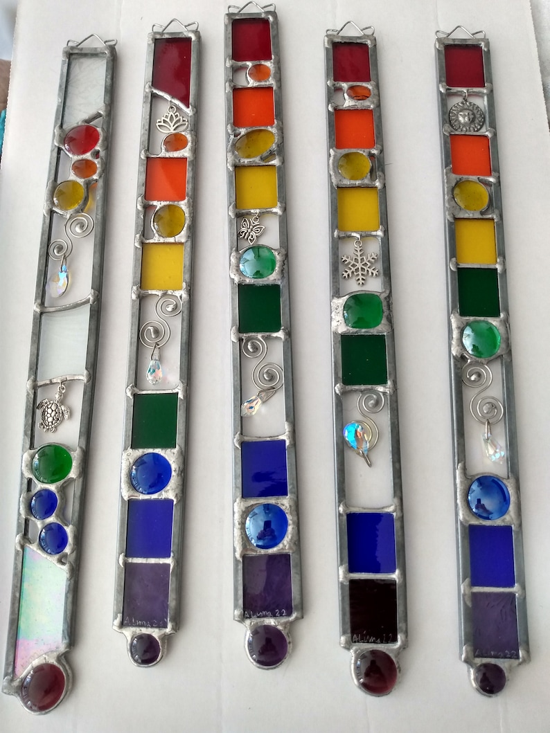 May include: Five stained glass suncatchers with a rainbow color scheme. Each suncatcher features a different design, including a snowflake, a butterfly, a turtle, a swirl, and a lion. The suncatchers are made with clear, red, orange, yellow, green, blue, and purple glass.