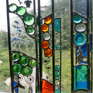 May include: A stained glass window hanging features a variety of colors and designs, including green, blue, orange, and white. The window is made up of several panels, each with a different design. The window is hanging in front of a window with a screen.