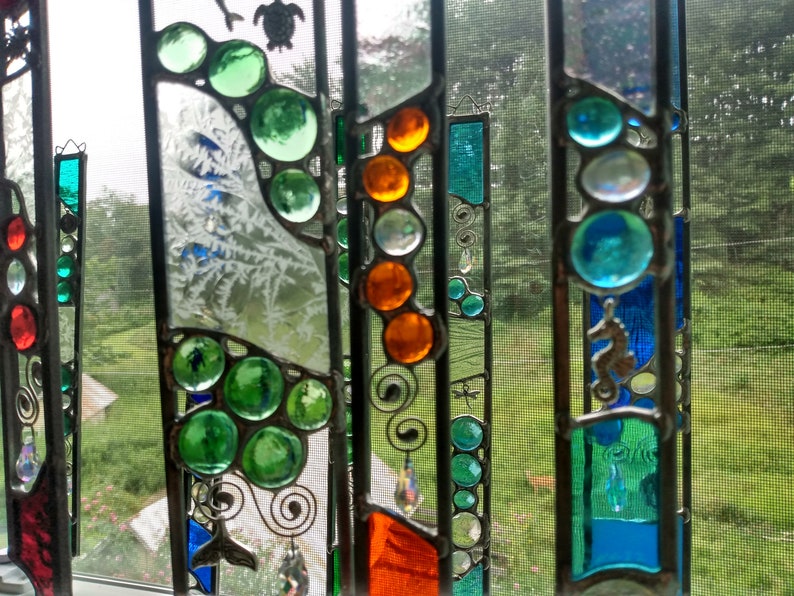 May include: A stained glass window hanging features a variety of colors and designs, including green, blue, orange, and white. The window is made up of several panels, each with a different design. The window is hanging in front of a window with a screen.