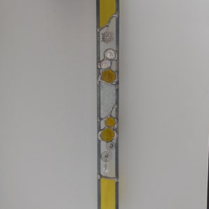May include: A stained glass window hanging with a yellow, white, and silver design. The design features a snowflake, circles, and swirls.