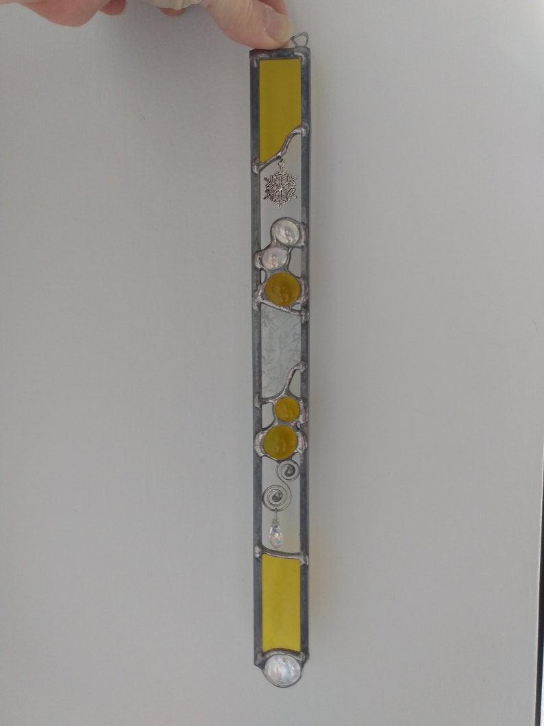 May include: A stained glass window hanging with a yellow, white, and silver design. The design features a snowflake, circles, and swirls.