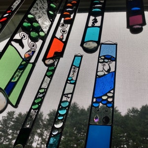 May include: A set of colorful stained glass suncatchers with abstract designs. The suncatchers are made of glass and feature various colors, including blue, green, and orange. They are hanging in a window and are illuminated by the sun.