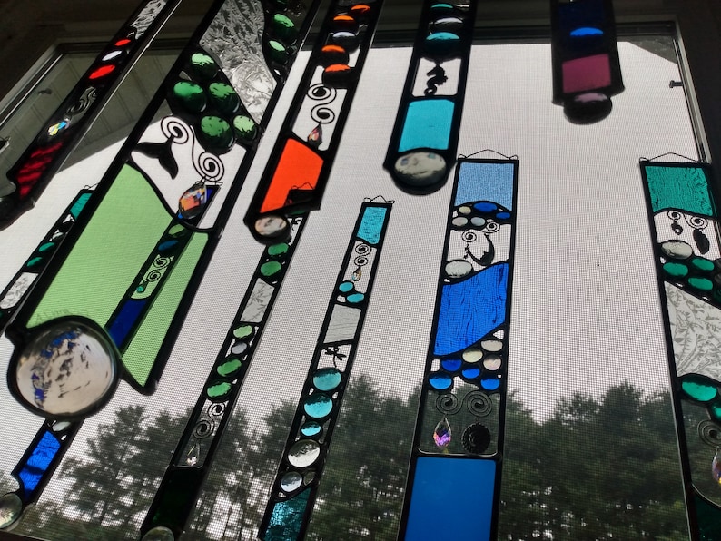 May include: A set of colorful stained glass suncatchers with abstract designs. The suncatchers are made of glass and feature various colors, including blue, green, and orange. They are hanging in a window and are illuminated by the sun.