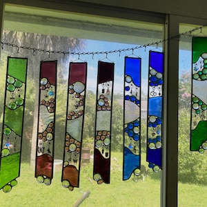 May include: A set of eight stained glass suncatchers hanging from a chain. The suncatchers are made of clear glass with colorful accents and feature a variety of designs, including circles, swirls, and geometric shapes.