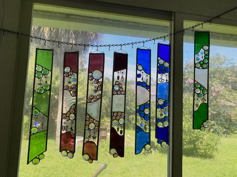 May include: A set of eight stained glass suncatchers hanging from a chain. The suncatchers are made of clear glass with colorful accents and feature a variety of designs, including circles, swirls, and geometric shapes.