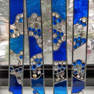 May include: Four stained glass panels with a blue and white color scheme. Each panel features a wavy design with round, clear glass accents. The panels are decorated with silver charms, including butterflies, stars, and swirls.