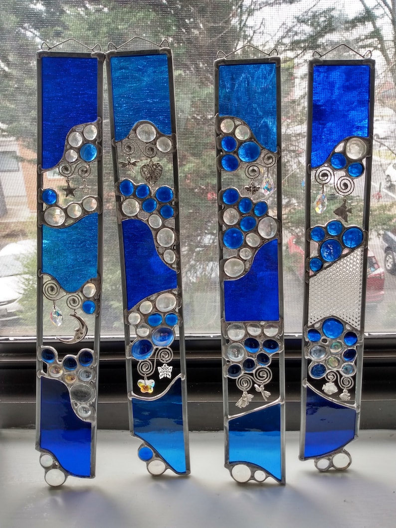 May include: Four stained glass panels with a blue and white color scheme. Each panel features a wavy design with round, clear glass accents. The panels are decorated with silver charms, including butterflies, stars, and swirls.