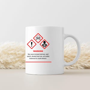 May include: White ceramic mug with a black and red warning label design. The label includes hazard symbols and the text "WARNING" with a list of potential side effects. The mug has a curved handle.