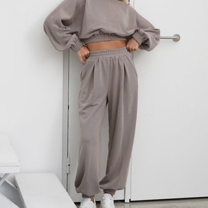May include: A two-piece outfit in a light brown color. The top is a cropped sweatshirt with long sleeves and a gathered elastic waistband. The pants are loose-fitting with a drawstring waistband and cuffed legs.