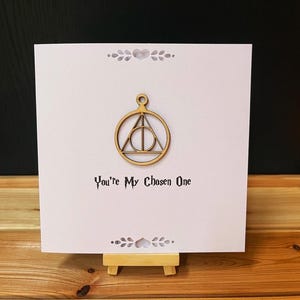 May include: A white greeting card with a wooden Deathly Hallows symbol and the text "You're My Chosen One." The card is displayed on a small wooden easel, with decorative floral accents.