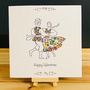 May include: A Valentine's Day card featuring a couple dancing in traditional folk attire. The card has a white background with a black outline drawing of the couple. Their clothing is decorated with colorful floral patterns, and the words "Happy Valentines" are written below.