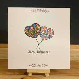May include: A Valentine's Day card featuring two heart-shaped balloons with floral patterns in various colors. The card has the words "Happy Valentines" written in black script. The card is displayed on a small wooden easel.