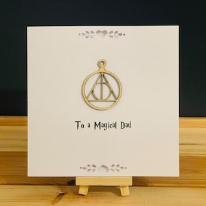 Può includere: Un biglietto color crema con la scritta "To a Magical Dad" in un carattere stilizzato. Il biglietto presenta un simbolo in legno dei Doni della Morte e ritagli decorativi. Il biglietto è esposto su un piccolo cavalletto di legno.