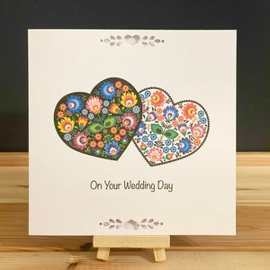 May include: A square wedding card featuring two heart shapes filled with colorful floral patterns. The card reads "On Your Wedding Day" and is displayed on a small wooden easel.
