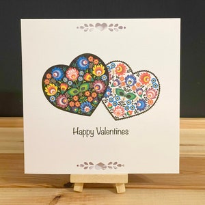 May include: A white card with two overlapping hearts decorated with colorful folk art designs. The text "Happy Valentines" is printed below the hearts.