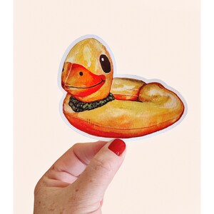 May include: A watercolor sticker of a yellow rubber duck with an orange beak and a green collar. The duck has a black eye and is smiling. The sticker is held by a hand with red nail polish.
