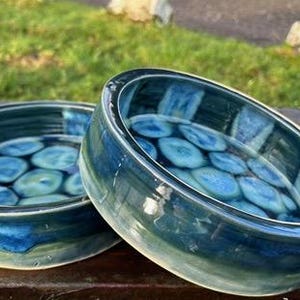 May include: Two handmade ceramic bowls with a glossy finish. The bowls are round with a dark blue exterior and a light blue and green interior design. The interior features a pattern of blue circles. The bowls are on a wooden surface.