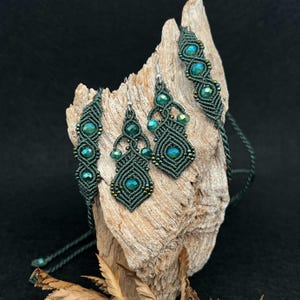 Può includere: Un set di gioielli in macramè verde scuro composto da collana, bracciale e orecchini. Il set include perline verde scuro e dettagli metallici. I gioielli sono esposti su un pezzo di legno trasportato dal mare.