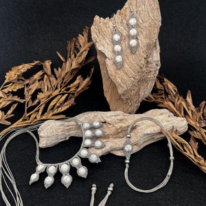 Può includere: Una collezione di gioielli fatti a mano con perline di pietra bianca e metallo argentato. Il set include una collana, orecchini e un braccialetto, esposti su un pezzo di legno trasportato dalla corrente su uno sfondo scuro. I gioielli hanno un'estetica bohémien.