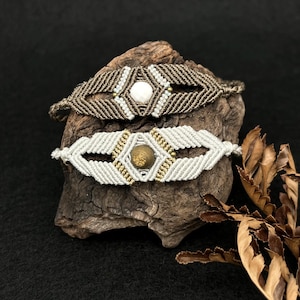 Può includere: Due braccialetti in macramè. Il braccialetto superiore è marrone con una pietra centrale bianca e dettagli bianchi e dorati. Il braccialetto inferiore è bianco con una pietra centrale dorata e dettagli dorati. Entrambi i braccialetti sono esposti su un pezzo di legno.