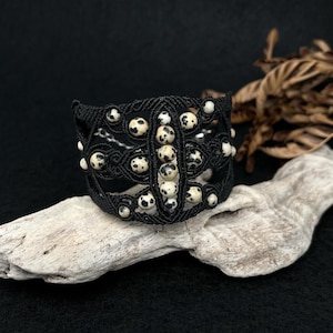 Könnte beinhalten: Ein schwarzes Makramee-Armband mit cremefarbenen, gesprenkelten Perlen. Das Armband hat ein offenes Design mit mehreren Reihen gewebtem Makramee und einer zentralen Reihe von Perlen. Die Perlen haben ein dalmatinerartiges Muster.