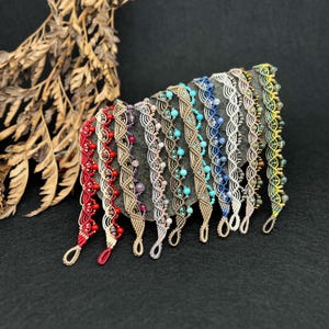 Può includere: Una collezione di braccialetti in macramè in vari colori, tra cui rosso, beige, turchese e blu, ognuno ornato di perline. I braccialetti sono esposti su una superficie grigio scuro, mostrando i loro intricati disegni intrecciati e le diverse palette di colori.