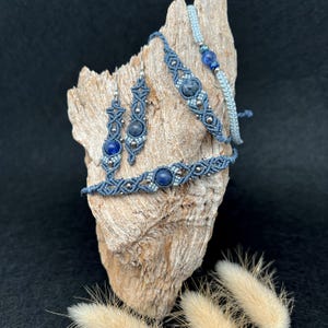 Può includere: Una collezione di gioielli fatti a mano, tra cui orecchini, bracciali e una collana, esposti su un pezzo di legno. I gioielli presentano cordoncino macramè blu e perline di pietre preziose blu, con dettagli color argento. Lo sfondo è nero.