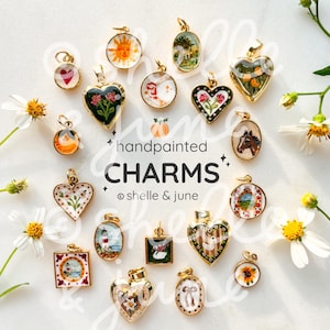 May include: A collection of small, gold-rimmed handpainted charms featuring various motifs including flowers, animals, landscapes, and celestial symbols. The text reads handpainted CHARMS © shelle & june.
