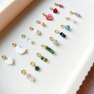 May include: A collection of small, decorative charms with gold-colored loops. The charms feature various shapes and colors, including hearts, stars, pearls, and gemstones in shades of pink, red, blue, and green. The charms are arranged on a white tray.