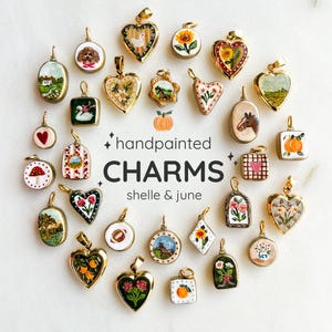 May include: A collection of hand-painted charms in various shapes, including hearts, ovals, and squares. The charms feature detailed miniature paintings of landscapes, flowers, animals, and other designs. The text "handpainted CHARMS shelle & june" is also visible.