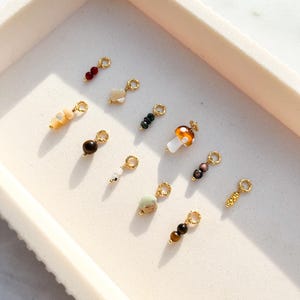 May include: A collection of small, decorative charms with gold-colored loops. The charms feature various natural stone beads in shades of red, brown, green, and white. One charm is shaped like a mushroom. The charms are arranged in a white tray.