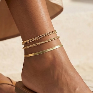 May include: Three gold anklets of different designs adorn an ankle, with a chain, rope, and flat band style. The person is wearing brown leather flip-flops. The image is taken from a low angle, showing the foot and lower leg.
