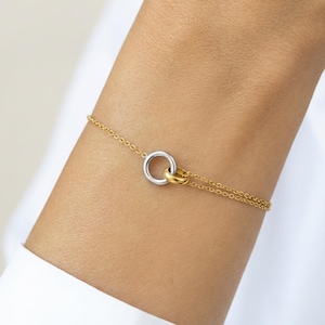 May include: A delicate gold-toned bracelet featuring a double-circle design. One circle is silver-toned, and the other is gold-toned, connected by a small gold-toned clasp. The bracelet is worn on a wrist, against a white shirt.