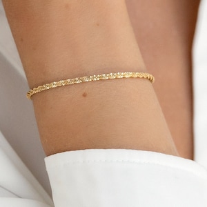 以下が含まれることがあります： テクスチャ加工されたツイストデザインの繊細なゴールドブレスレット。白いシャツを背景に、腕に着用されています。このジュエリーはシンプルでありながらエレガントなアクセサリーです。