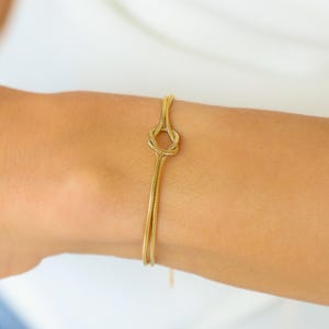 Puede incluir: Un brazalete dorado con un diseño de nudo. El brazalete está hecho de metal fino y flexible y se usa en la muñeca. El diseño es minimalista y elegante, adecuado para el uso diario.